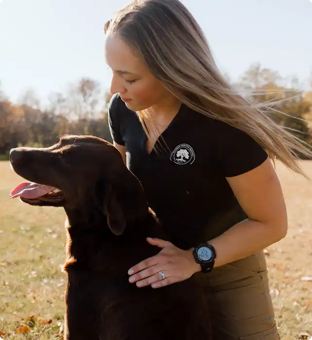 Woman practitioner in outdoor field cradling calm black dog with relaxed expression, demonstrating compassionate somatic care and connection